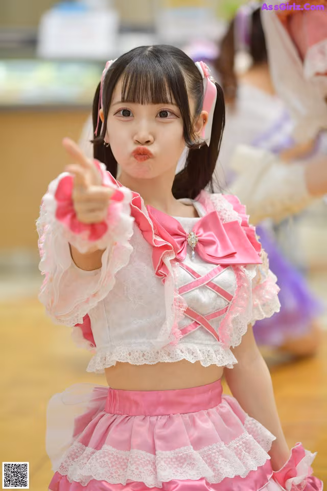 A young girl in a pink and white outfit pointing at the camera.